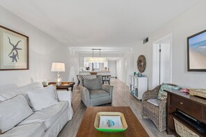 Una sala de estar con sofá, sillón y mesa de centro. Se observa una cocina al fondo con zona de comedor.