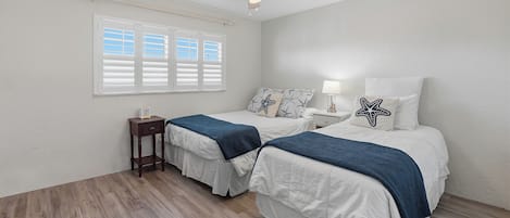A bedroom with two beds, a ceiling fan, a window with blinds, and a bedside table with a lamp.