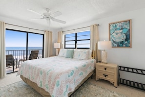 A bedroom with a bed, bedside tables, a ceiling fan, a painting, and a view of the ocean.