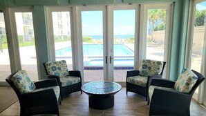 Une pièce comprenant quatre chaises en osier avec des coussins à motifs floraux, une table basse centrale et de grandes portes vitrées donnant sur un espace piscine.