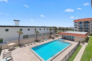 A swimming pool with lounge chairs and umbrellas, surrounded by a building with a red roof and a white facade. - 310 - Incredible ocean and pool views 1 bedroom (New Smyrna Beach)