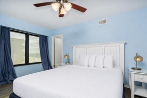Une chambre à coucher avec un grand lit, un ventilateur de plafond, deux tables de chevet avec des lampes, une fenêtre avec des rideaux et une porte.