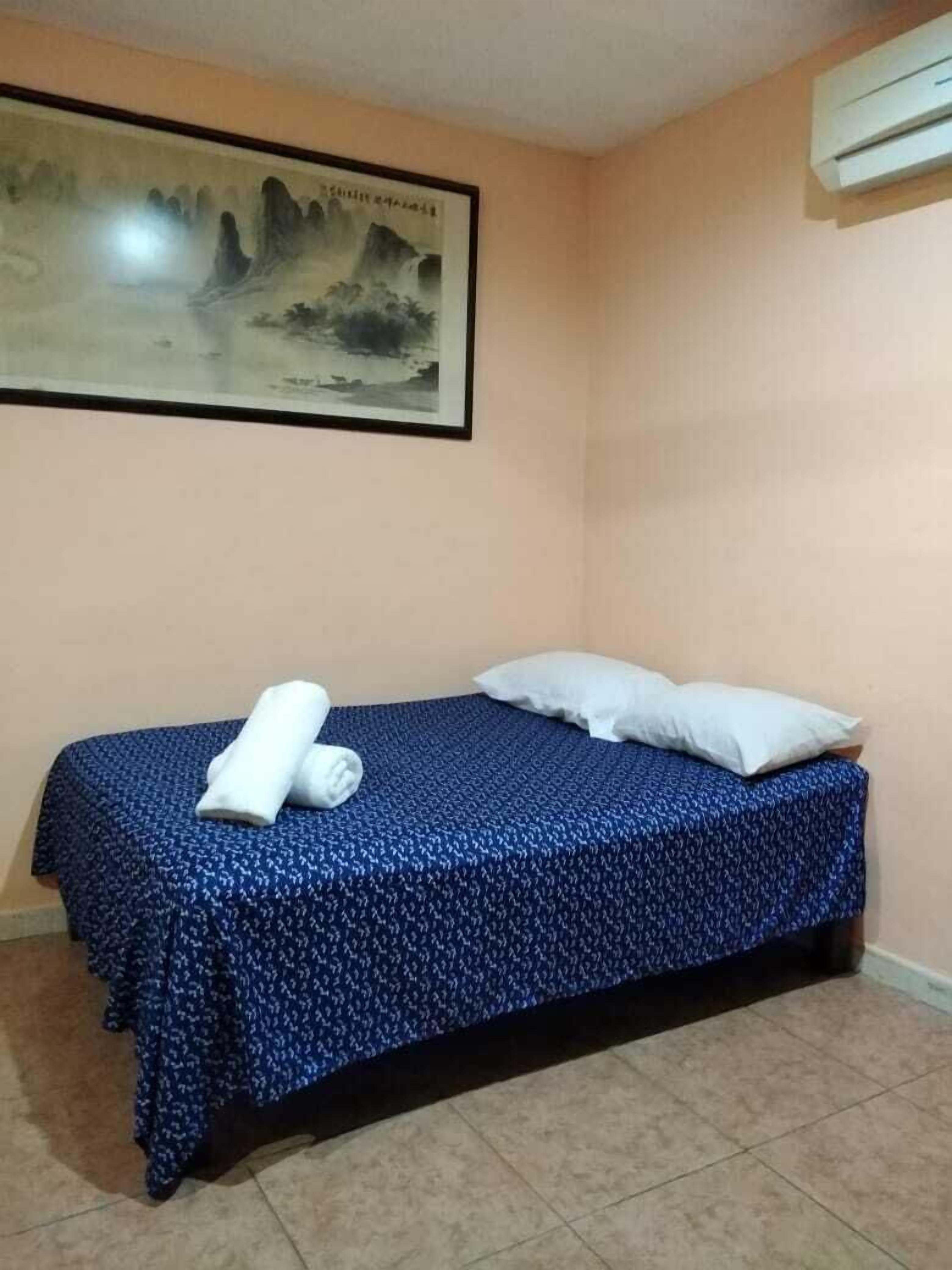 A single bed with a blue patterned bedspread, two pillows, and a rolled white towel on the bed.