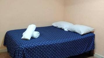 A single bed with a blue patterned bedspread, two pillows, and a rolled white towel on the bed.