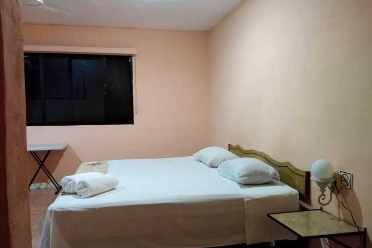 A single bed with white linens, a bedside table with a lamp, and a window with blinds.