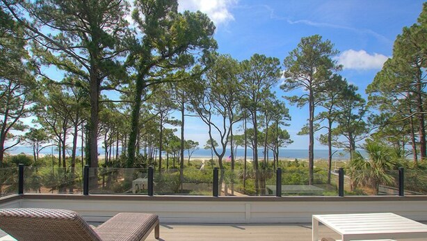 Une terrasse dotée de deux chaises longues et d’une table, offrant une vue sur une plage bordée de grands arbres et sur l’océan.