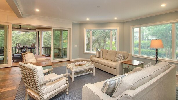 A spacious living room with a sofa, armchair, and coffee table, large windows, and a sliding glass door leading to an outdoor area.