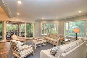 A spacious living room with a sofa, armchair, and coffee table, large windows, and a sliding glass door leading to an outdoor area.