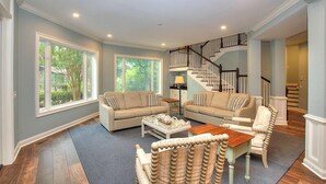 A spacious living room with two sofas, a coffee table, and a wicker chair. A staircase with a black railing leads to an upper level.