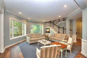 A spacious living room with two sofas, a coffee table, and a wicker chair. A staircase with a black railing leads to an upper level.