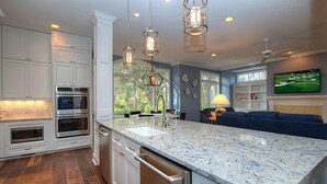 A modern kitchen with white cabinetry, granite countertops, and stainless steel appliances. There is a large island with a sink and a dining area with a blue sofa and a TV mounted on the wall.