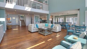 A spacious living area with a high ceiling, a balcony, and a dining area with blue furniture. - 123 Dune Ln - Oceanfront Home in N. Forest Beach w/ Private Pool & Beach Path (Hilton Head Island)