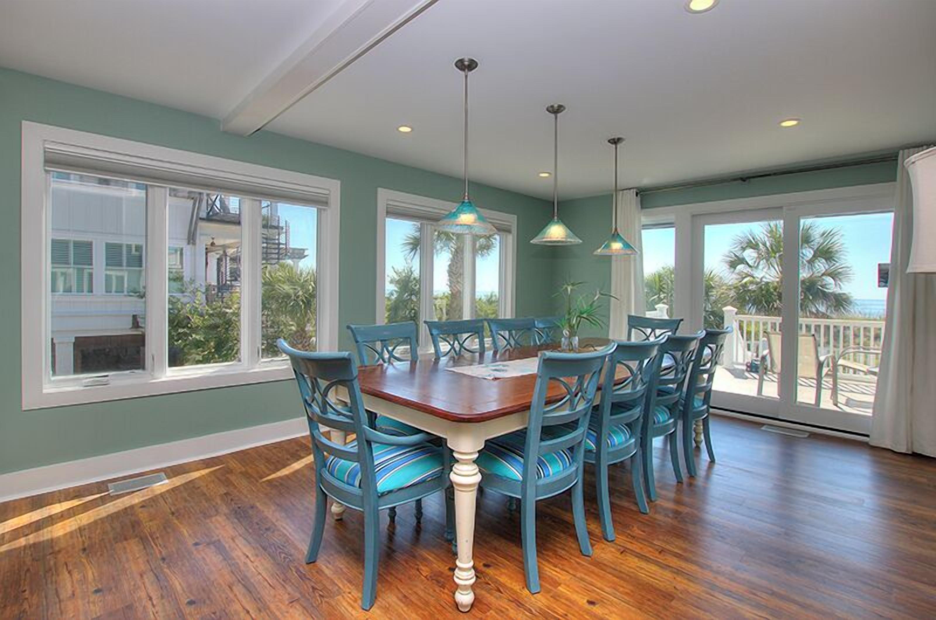 Un comedor con mesa de madera y sillas azules, ventanas amplias con vistas a palmeras y un edificio, y lámparas colgantes.