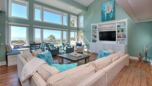 A spacious living room with large windows, a comfortable seating area, a flat-screen TV, and a bookshelf. - 123 Dune Ln - Oceanfront Home in N. Forest Beach w/ Private Pool & Beach Path (Hilton Head Island)