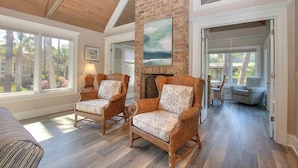 A spacious living room with wicker chairs, a brick fireplace, and large windows offering a view of palm trees. - 26 Surf Scoter - 2nd Row Luxury Home in Sea Pines w/ Private Pool & Spa (Hilton Head Island)