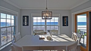 Un comedor con una mesa grande, sillas, lámpara colgante y ventanas amplias con vista a la playa.