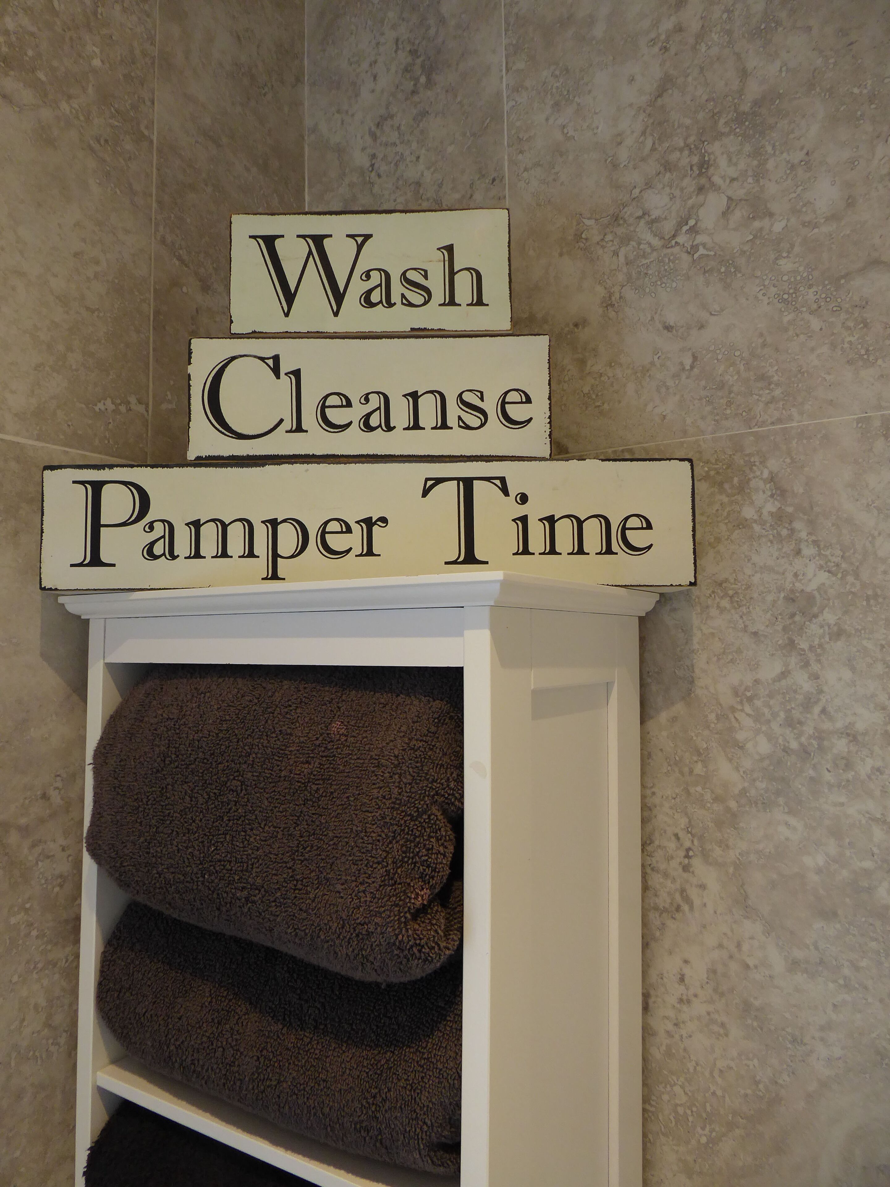 A white shelf with three signs stacked on top, reading 'Wash,' 'Cleanse,' and 'Pamper Time.' Below the shelf, there are folded brown towels.