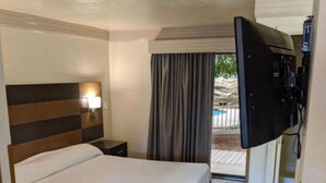 A hotel room with a bed, a nightstand, a TV, and a view of the outdoors. - Atrium Inn (Fort Stockton)