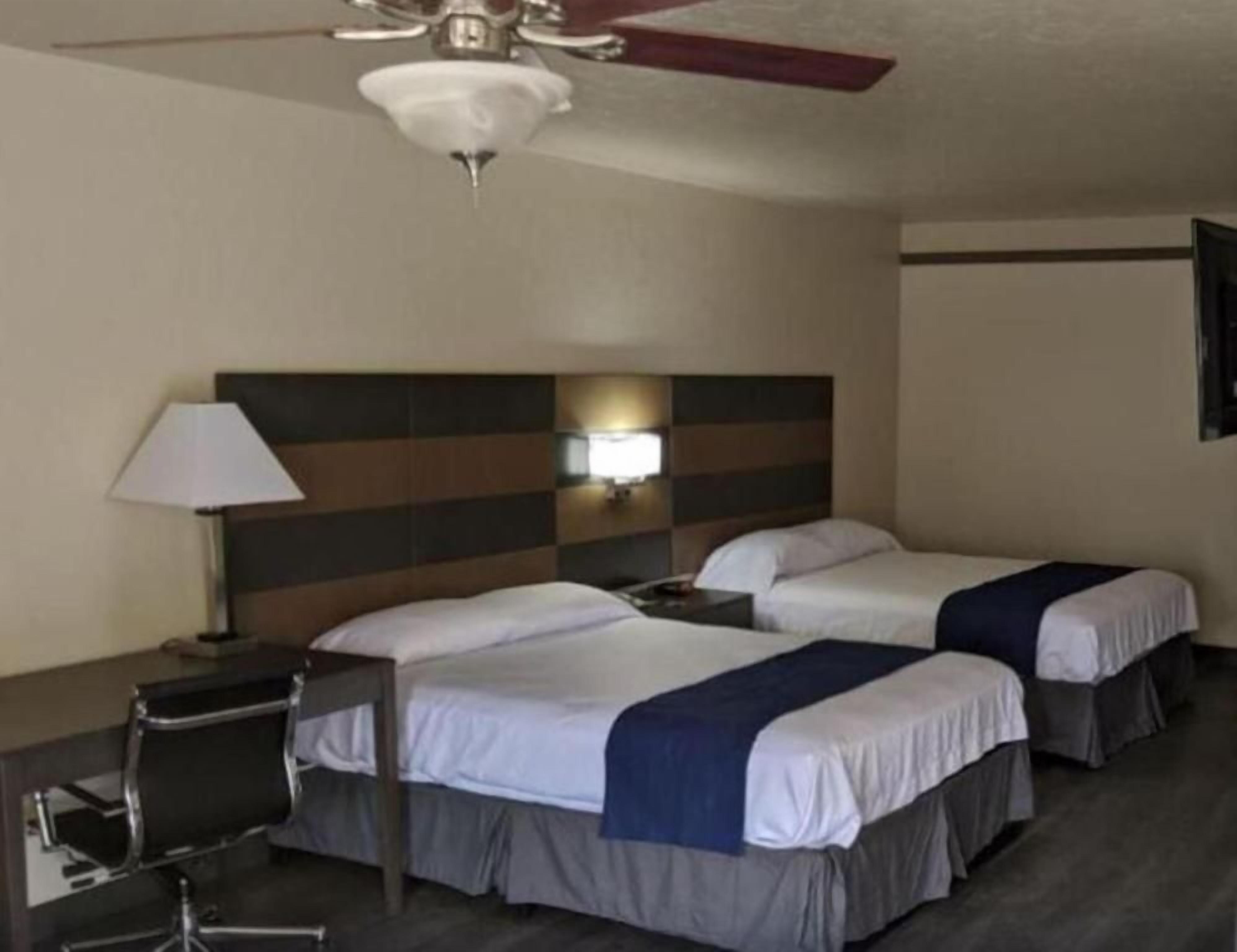 A hotel room with two beds, a desk, a chair, a lamp, and a ceiling fan.