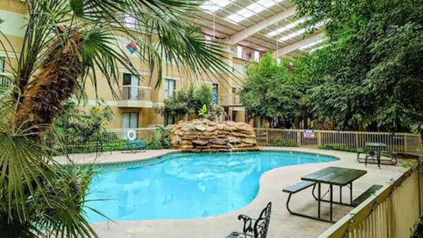 An indoor swimming pool with a stone feature, surrounded by greenery and a covered area with seating. - Atrium Inn (Fort Stockton)