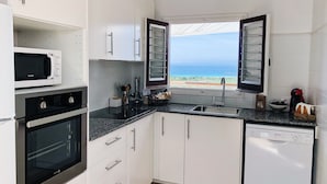 A modern kitchen with white cabinets, a microwave, oven, and dishwasher. A window offers a view of the ocean. - Villa Maria, incredible sea views, relax, swimming pool, wifi, barbecue, garden (alaior)