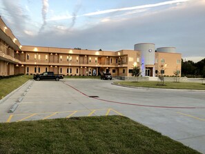 A multi-story hotel with a parking lot, a building with a cylindrical structure, and a landscaped area with small trees. - Crystal Inn (Houston)