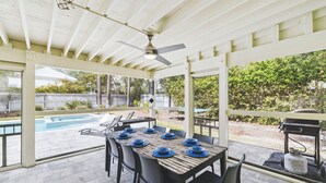 Un patio exterior con una mesa dispuesta para comer, un ventilador de techo y vista a un jardín trasero.