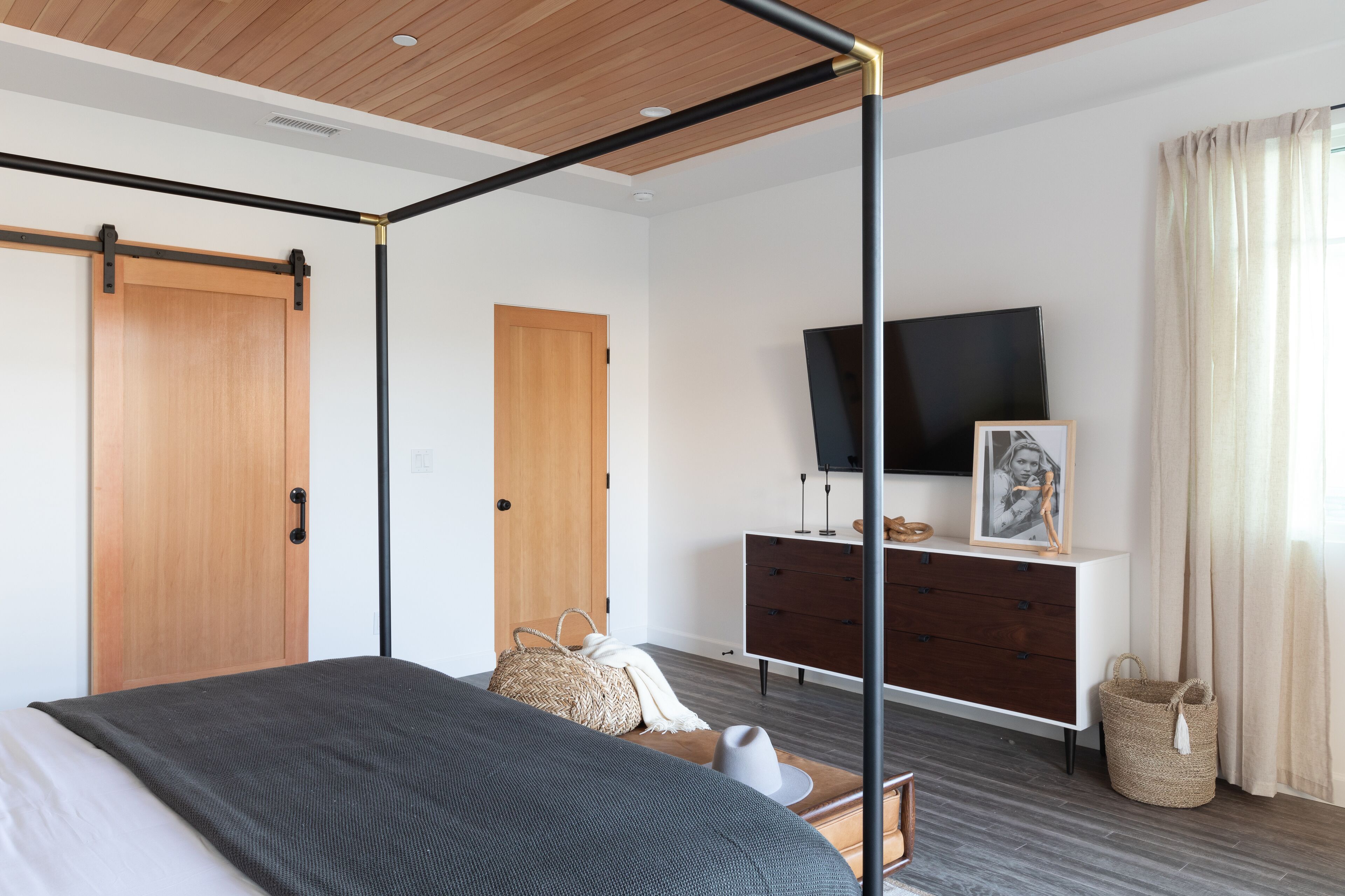 A modern bedroom with a four-poster bed, wooden doors, a TV, a dresser, and a large window with curtains.