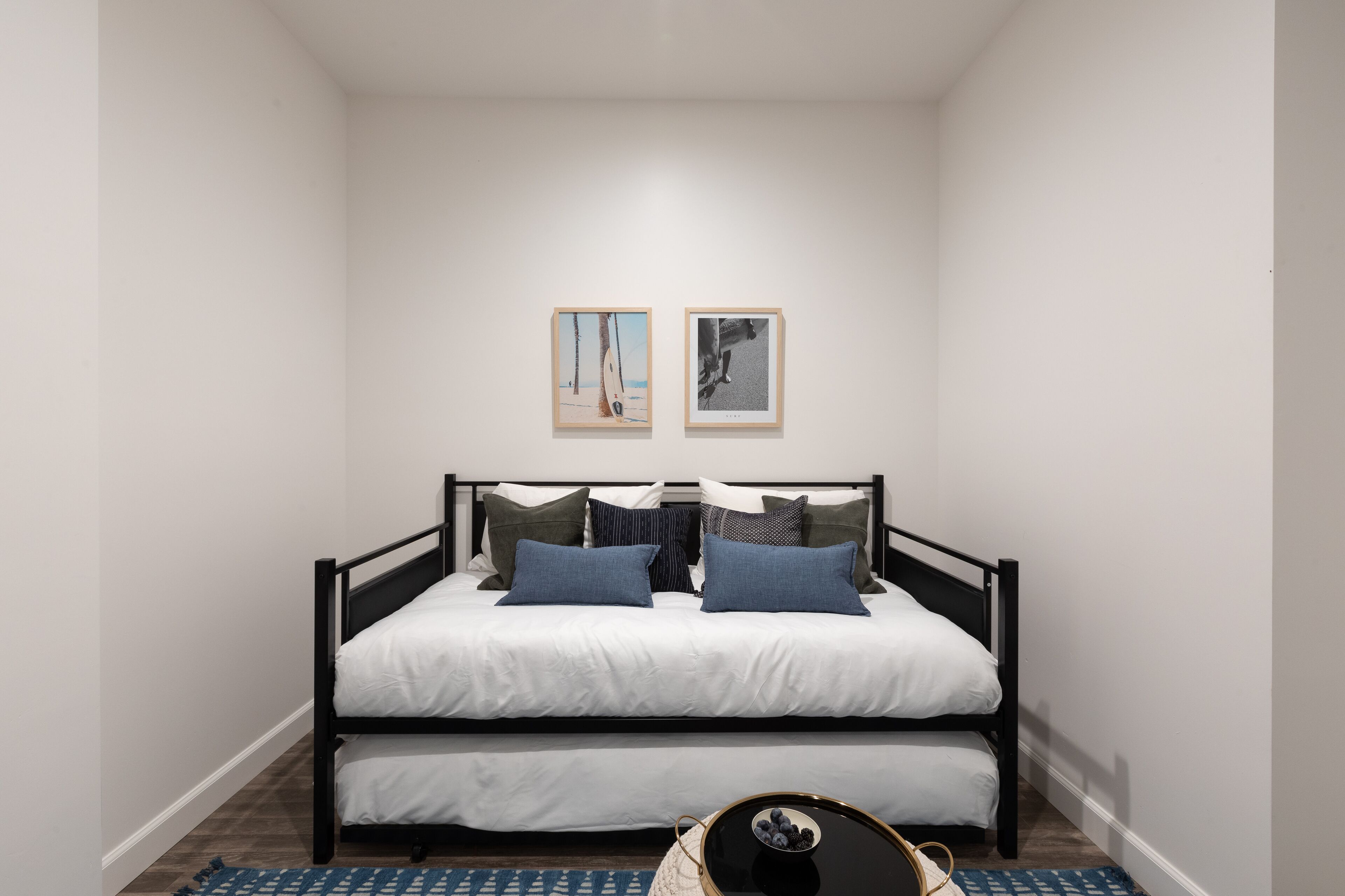 Ein Schlafzimmer mit schwarzem Metallbettgestell, weißer Bettwäsche und einem blau gemusterten Teppich. An der Wand hängen zwei gerahmte Bilder.