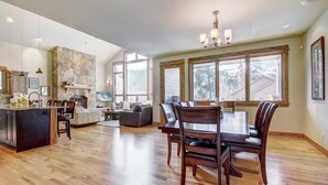 A spacious kitchen with a stone accent wall, a dining area with a wooden table and chairs, and large windows offering a view of the outdoors. - Riverwood Retreat-4BR Warrior's Mark Home, Private Hot Tub, Fireplace, Sleeps 12 (Breckenridge)