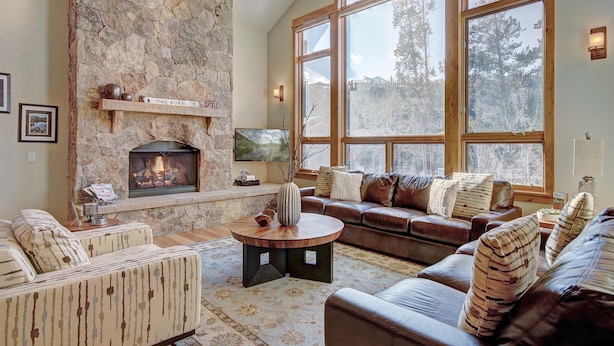 A cozy living room with a stone fireplace, leather sofas, and large windows offering a view of trees. - Riverwood Retreat-4BR Warrior's Mark Home, Private Hot Tub, Fireplace, Sleeps 12 (Breckenridge)