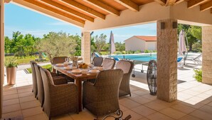 Un área de comedor al aire libre techada con mobiliario de mimbre, una piscina y vistas del paisaje circundante.