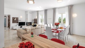 Una sala moderna con sofá, televisor y chimenea. Hay un comedor con una mesa preparada para dos personas y vistas al exterior.
