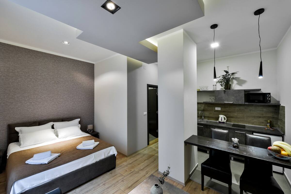 A modern hotel room with a bed, bedside table, and a kitchenette area.