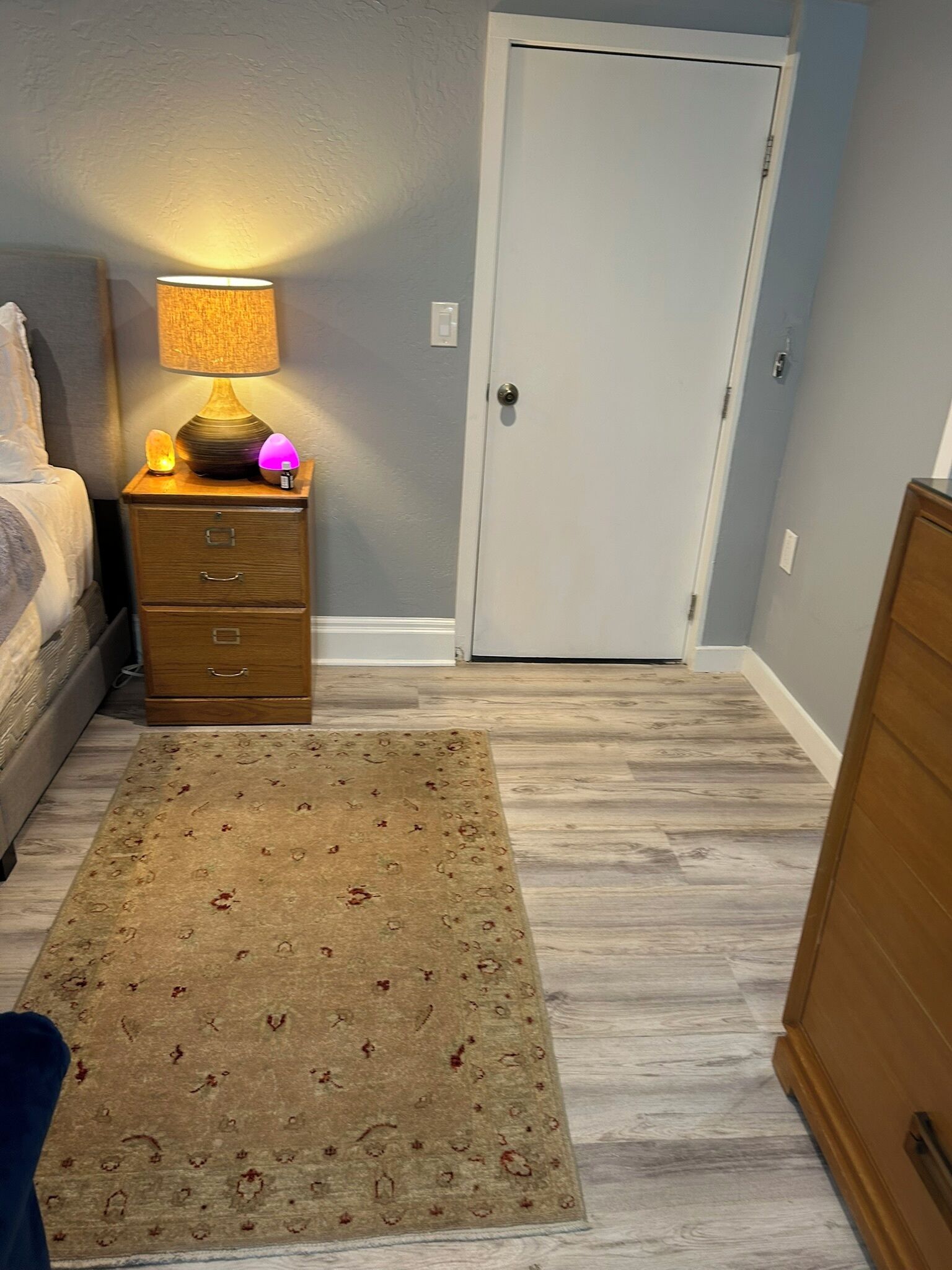 A bedroom with a bed, a nightstand with a lamp, a wooden dresser, and a patterned rug on a wooden floor.