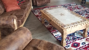 Un salón con un sofá de cuero marrón, un sillón a juego, una mesa de centro de madera y una alfombra estampada. Hay armarios de madera y una puerta con una cruz en la pared.