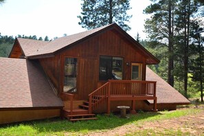 Situated in a forested area, this cabin features a front porch with a bench and a small table, and a set of stairs leading to the entrance.