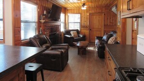 A cozy cabin interior with a fireplace, leather seating, a kitchen area, and wooden walls. - 2+2, Spa, Sleeps 6, Walk 2 Hiking & Biking Trails, Pet Friendly on North Shore (Big Bear)