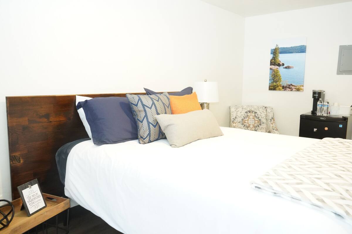 A neatly made bed with a wooden headboard, multiple pillows, a bedside table with a lamp, a framed picture on the wall, and a small nightstand with a coffee maker.