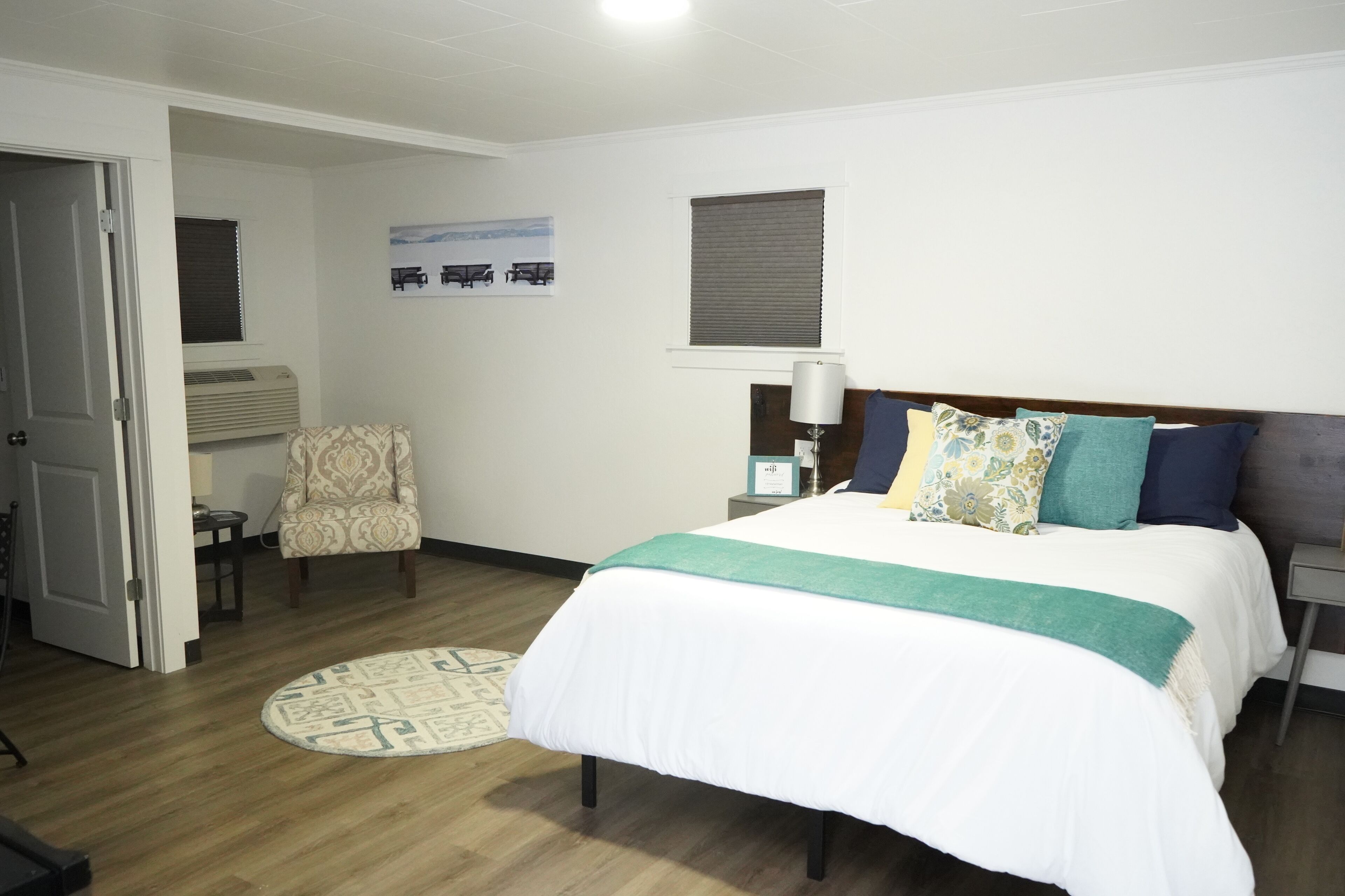 Quarto de hotel com cama, poltrona, mesa pequena, luminária e janela com persianas.