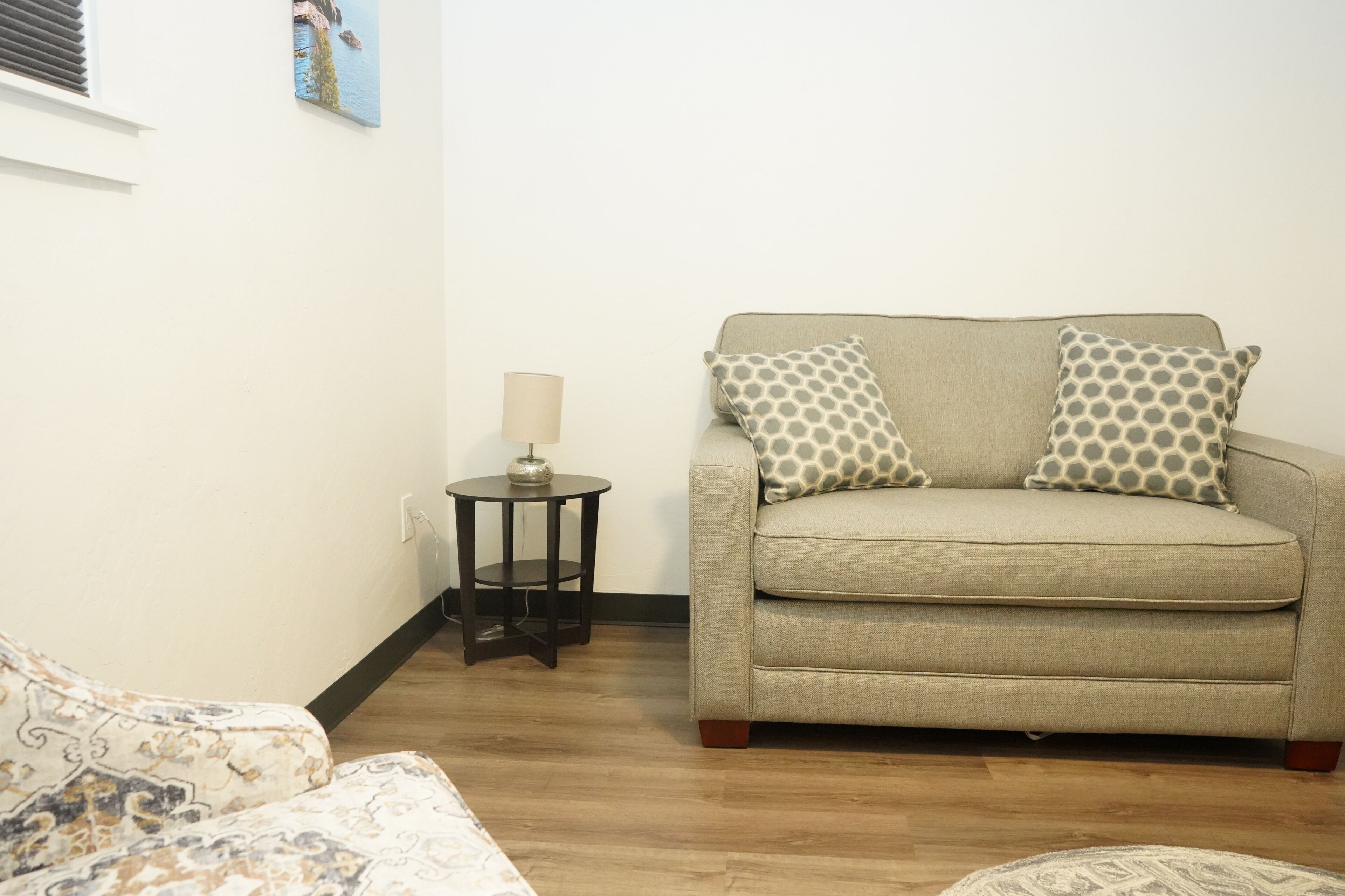 A living room with a beige sofa, patterned cushions, a small round table with a lamp, and a wooden floor.