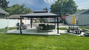 A covered patio area with picnic tables and a grill, surrounded by a white fence and a grassy lawn. - North Idaho Inn (Coeur d'Alene)