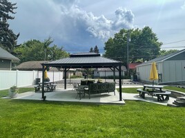 Un patio techado con mesas de picnic y una parrilla, rodeado por una cerca blanca y un césped.