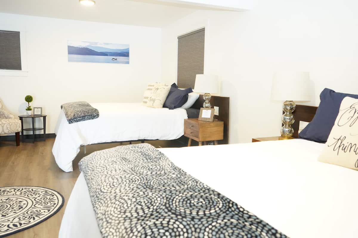 A hotel room with two beds, a bedside table with a lamp, a small side table with a plant, and a picture on the wall.
