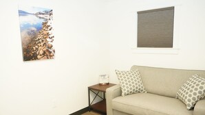 A living room with a beige sofa, patterned pillows, a small wooden side table, and a framed picture of a rocky shoreline. - North Idaho Inn (Coeur d'Alene)