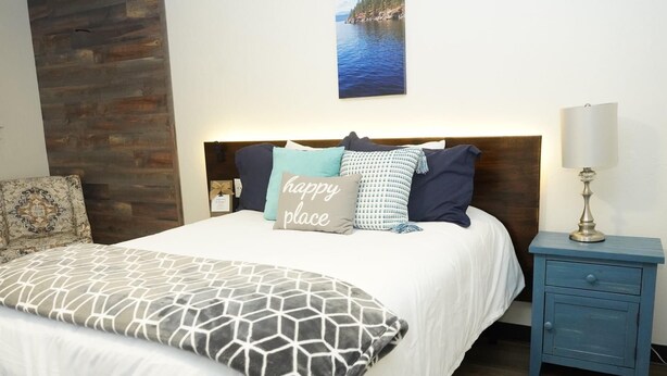 A neatly made bed with a white comforter and a geometric-patterned pillow, a blue nightstand with a lamp, and a wooden accent wall. - North Idaho Inn (Coeur d'Alene)