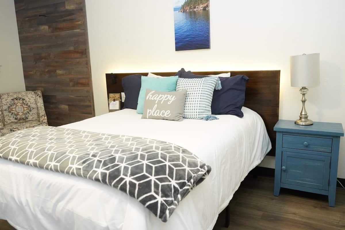 A neatly made bed with a white comforter and a geometric-patterned pillow, a blue nightstand with a lamp, and a wooden accent wall.