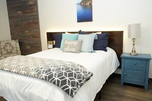 Een net opgemaakt bed met een witte dekbedovertrek en een kussen met een geometrisch patroon, een nachtkastje in blauw met een lamp, en een houten accentmuur.