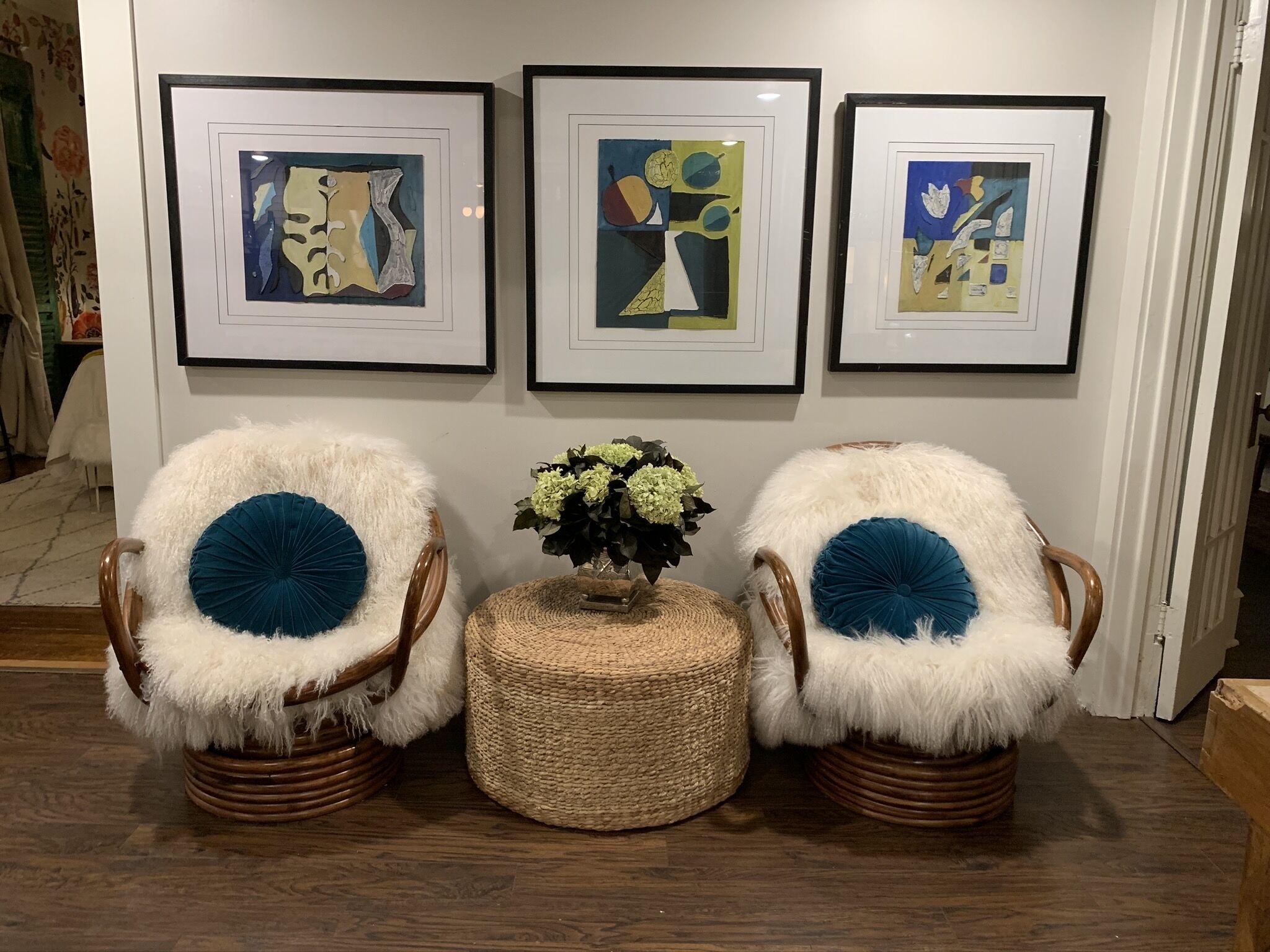 Une pièce décorée avec trois tableaux abstraits encadrés accrochés au mur, deux fauteuils moelleux dotés de coussins bleus, et un petit pouf en osier sur lequel est posé un vase rempli de fleurs.