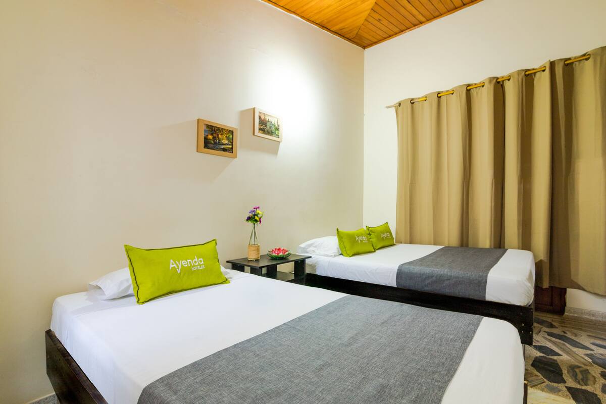 Une chambre d’hôtel avec deux lits, chacun recouvert d’une couverture grise et agrémenté d’un oreiller vert. On y trouve une petite table sur laquelle est posé un vase de fleurs, ainsi qu’un tableau accroché au mur.